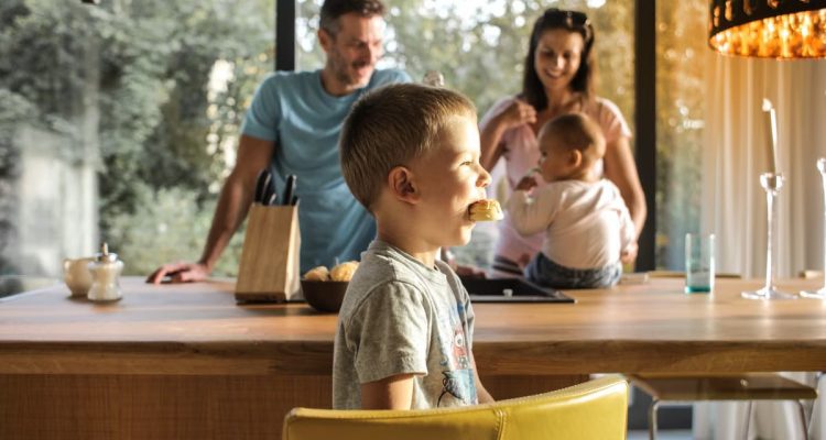 Side view of playful boy eating cookie while sitting at table with parents and sibling on background spending time at home