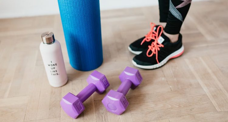 Unrecognizable sportswoman wearing sneakers and black leggings standing with sport mat on wooden floor near dumbbells and water bottle before exercising indoors
